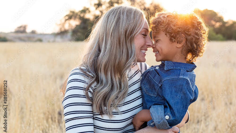 Beautiful happy Mother holding her adorable diverse son in the outdoor sunlight. Sun rays shining through as the mom shows love and affection to her son